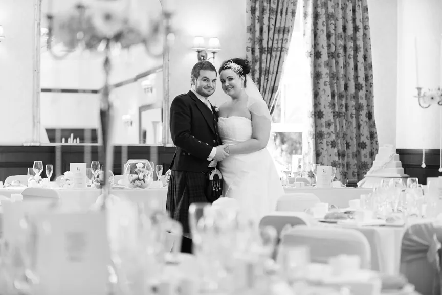 Bride in a white gown and groom in a traditional kilt suit posing together in a decorated wedding reception room.