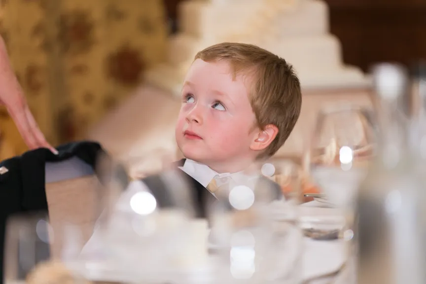 Young boy in formal attire looking up while seated at a dining table with blurred glassware in the foreground.