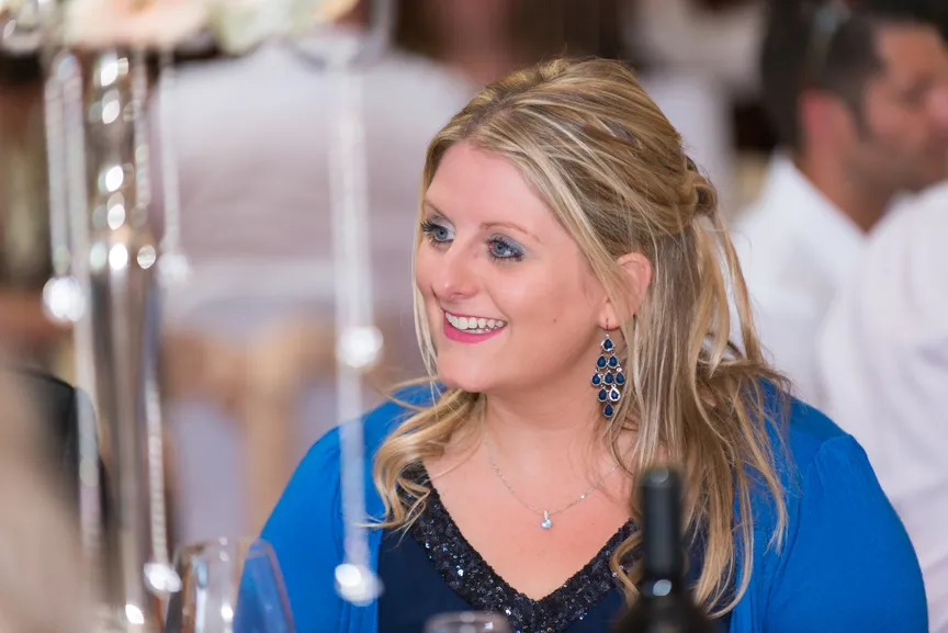 Smiling blonde woman with long hair wearing blue earrings and a blue outfit at a social event.