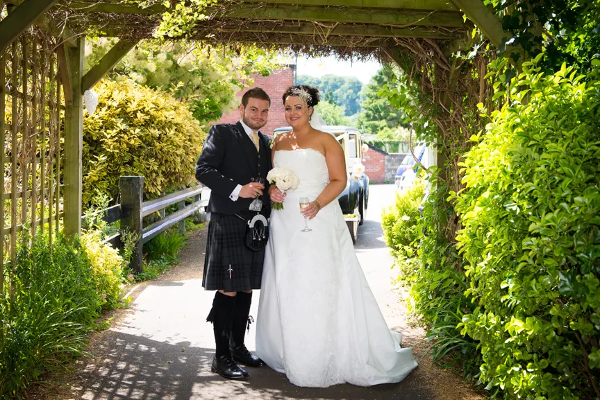 Bride in a white wedding dress and groom in traditional Scottish kilt standing under a foliage arch, holding drinks and smiling.