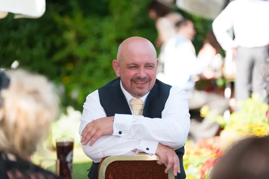 Smiling bald man in white shirt, gold tie, and dark vest sitting outdoors with arms crossed on the back of a chair.