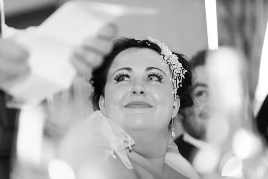 Black and white close-up of a bride with decorative hairpiece looking upward and smiling, with a blurred figure in the background.