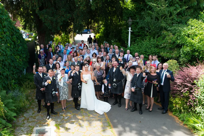 Large wedding group photo with bride in white gown, groom and male guests in kilts, and family and friends gathered outdoors.