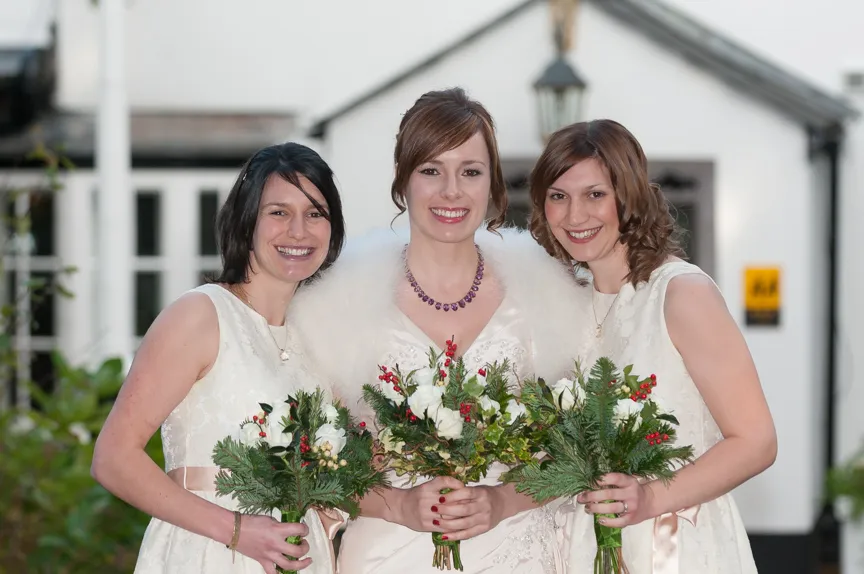 Three smiling women in white dresses holding bouquets with greenery and red berries, standing close together outdoors.