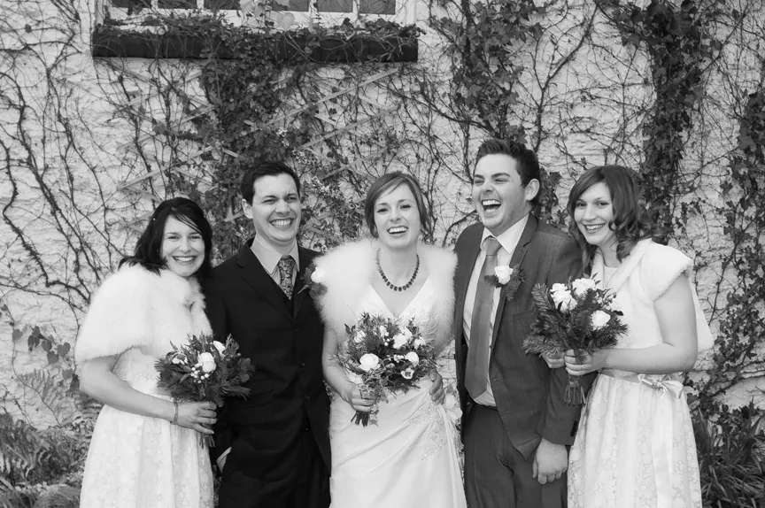 Black and white photo of a wedding party with three women in white dresses holding bouquets and two men in suits smiling and laughing in front of a wall covered in climbing vines.