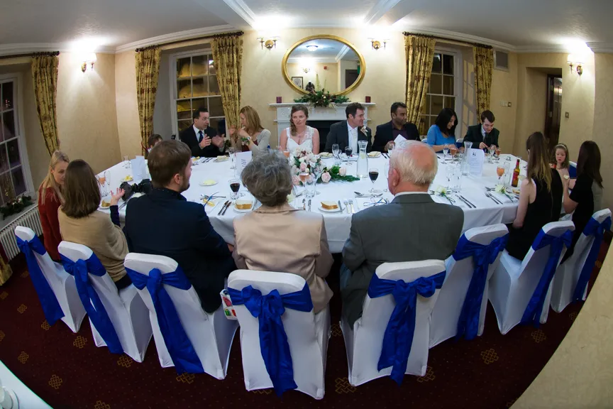 Guests seated around a decorated table with white covers and blue bows at a formal indoor event.