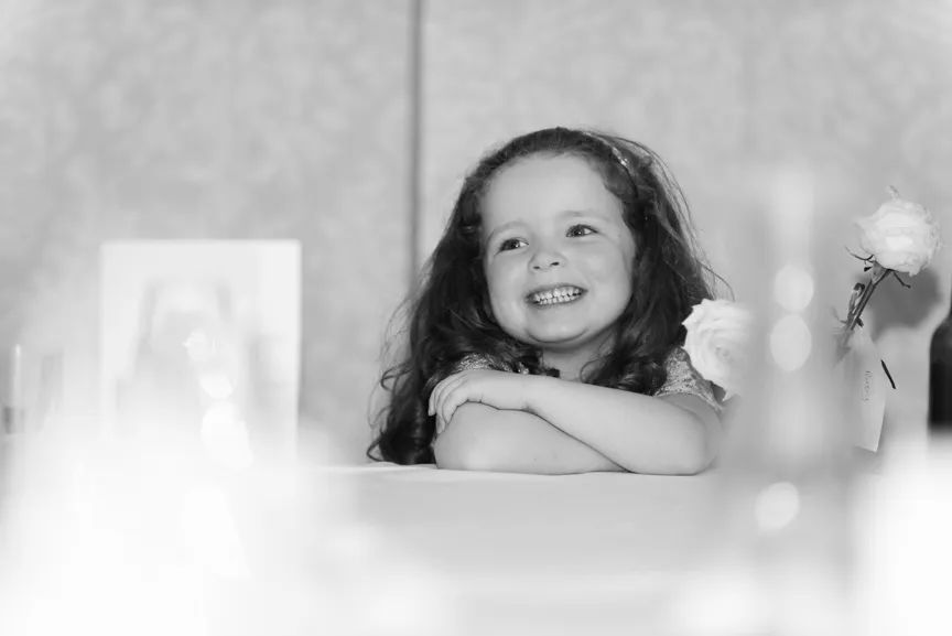 Smiling young girl with long curly hair resting her crossed arms on a table with a rose next to her.