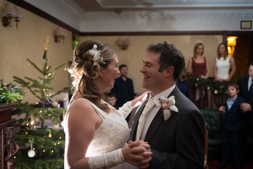Bride and groom smiling and dancing indoors near a decorated Christmas tree during a wedding celebration.
