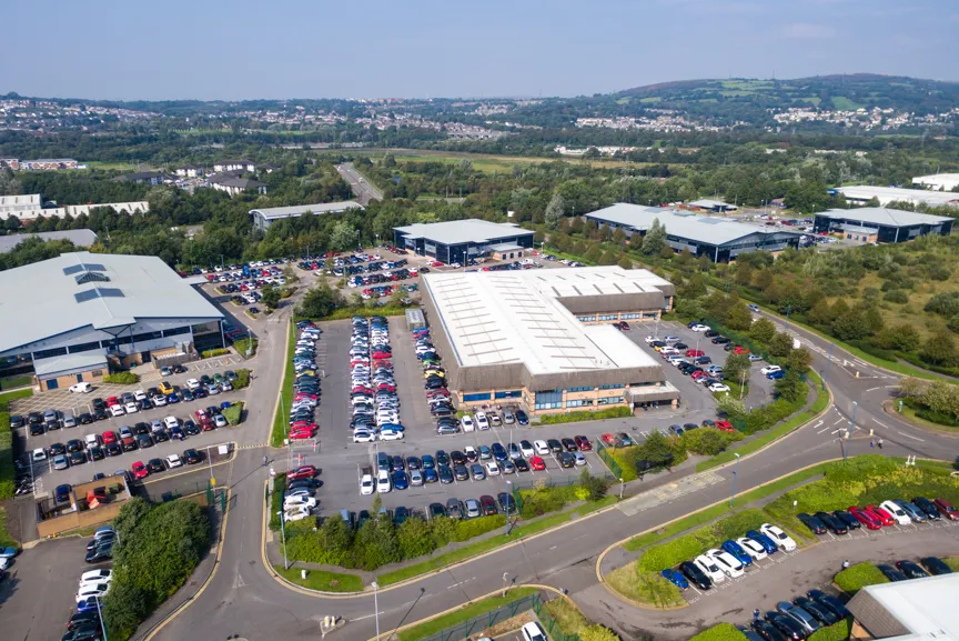 Aerial view of a commercial or industrial business park with multiple buildings and parking lots filled with cars surrounded by greenery and distant hills.