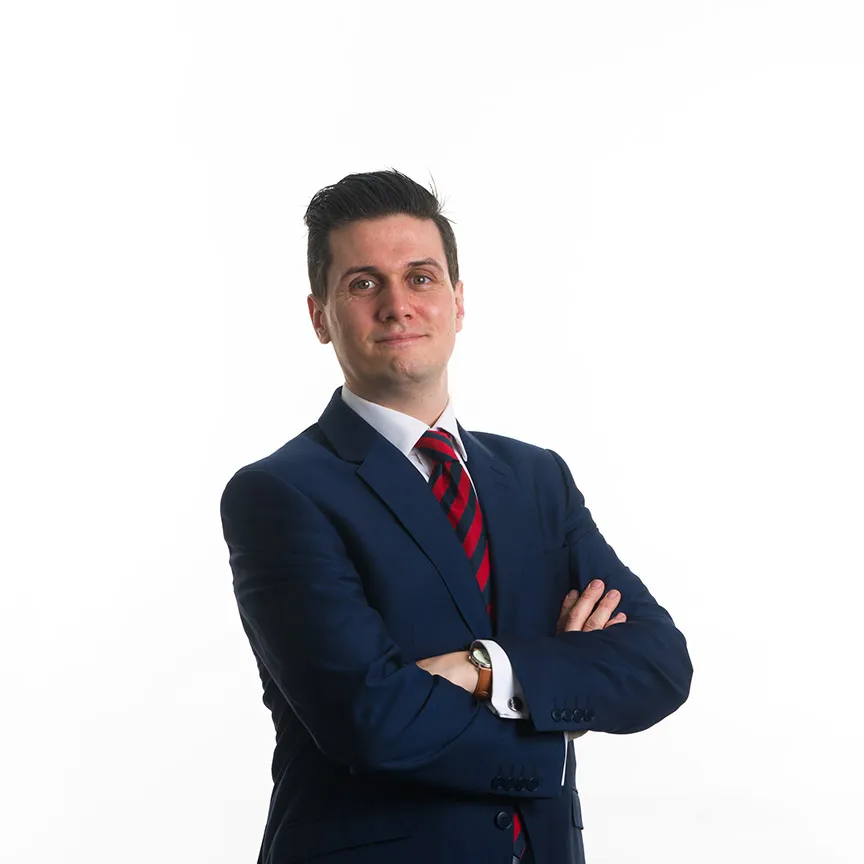 Man in a dark blue suit with a white shirt and red striped tie, standing with arms crossed.