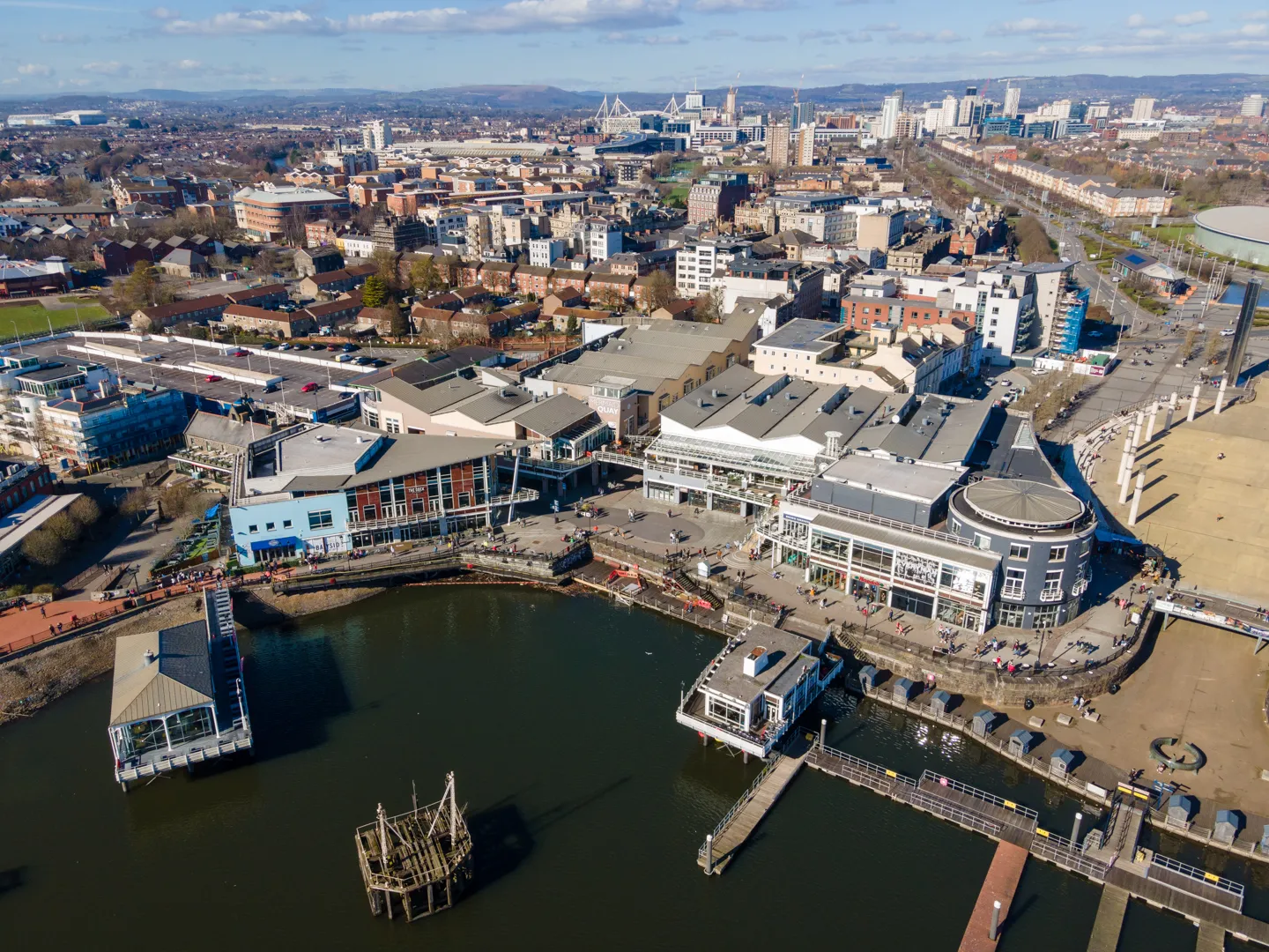 Aerial view of a waterfront shopping and entertainment area with a dock, restaurants, and nearby city buildings under a partly cloudy sky.
