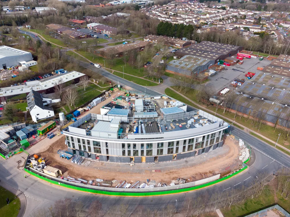 Aerial view of a curved, multi-story building under construction surrounded by roads and green fencing, with residential and industrial buildings in the background.