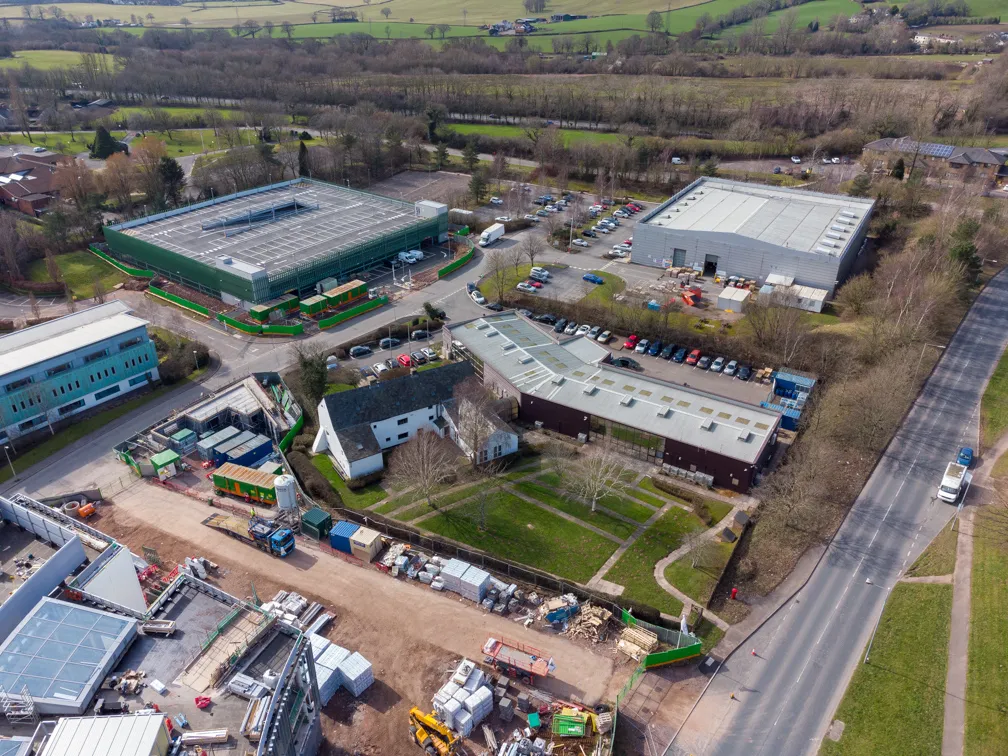 Aerial view of a construction site with several buildings, parked cars, and green fields in the background.