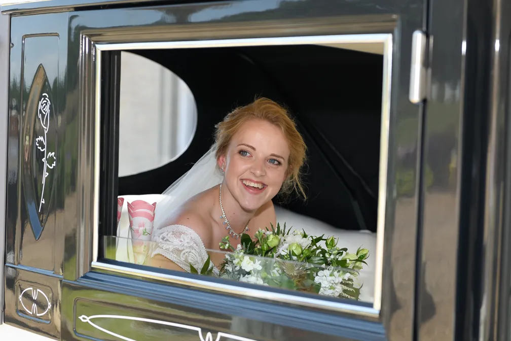 Smiling bride in a white dress holding a bouquet of white flowers, sitting inside a black vintage car.