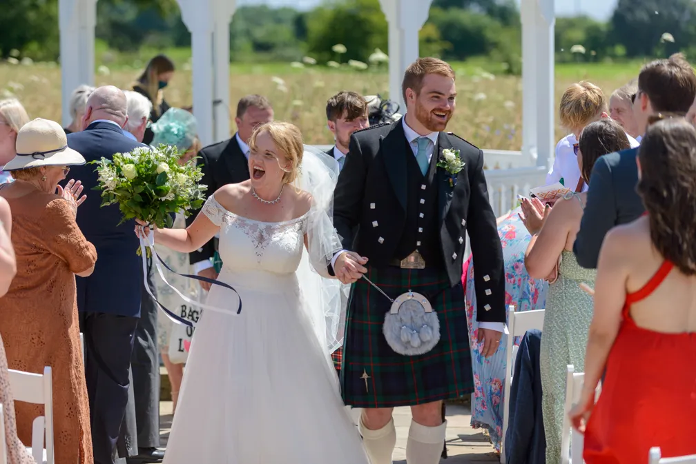 Bride in a white dress and groom in a traditional Scottish kilt holding hands and smiling while walking down an outdoor aisle with guests applauding.