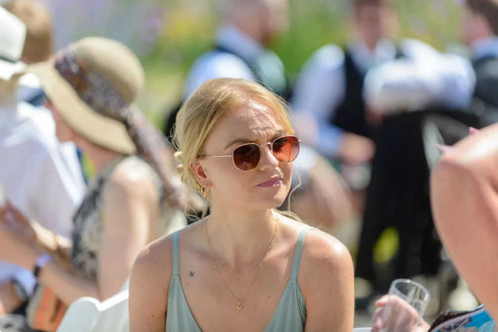 Blonde woman wearing sunglasses and a light green tank top at an outdoor event, surrounded by blurred people.