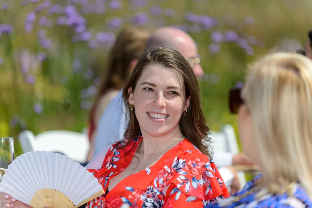 Smiling woman in a red floral dress holding a white hand fan while sitting outdoors at an event.