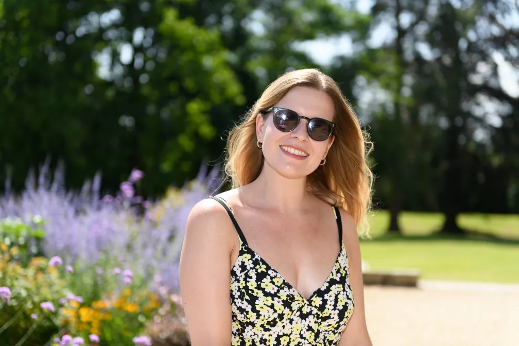 Smiling woman with blonde hair wearing sunglasses and a black floral dress outdoors with green trees and purple flowers in the background.
