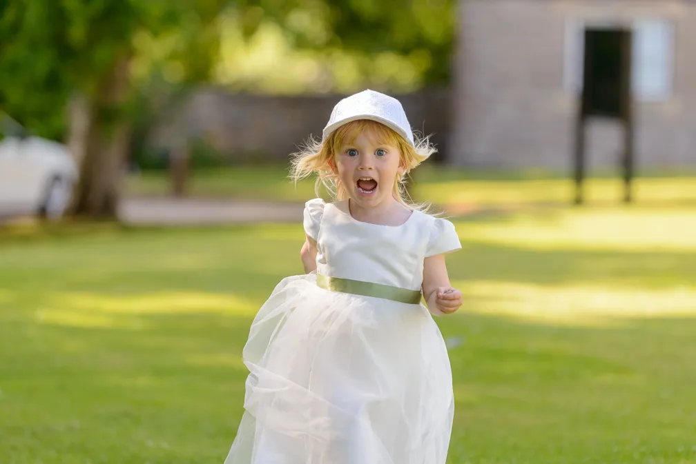 Young girl in a white dress and cap running on green grass with an excited expression.
