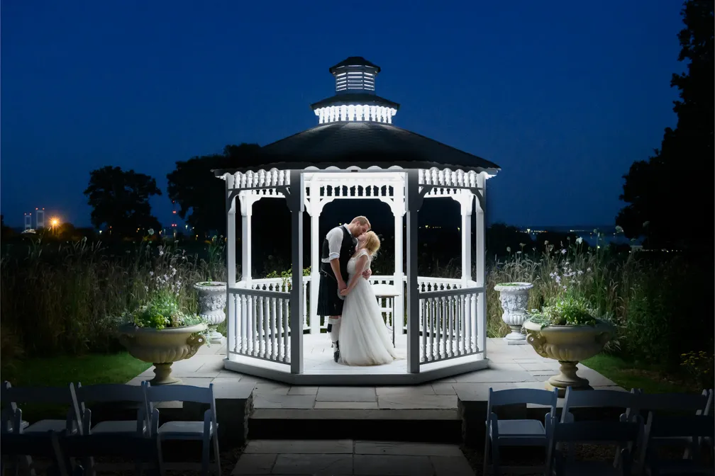 Bride and groom sharing a romantic moment inside a brightly lit white gazebo at night.