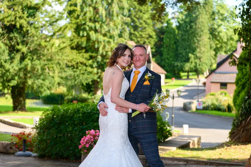 Bride in white lace dress and groom in checkered suit with orange tie embracing outdoors with greenery and buildings in the background.