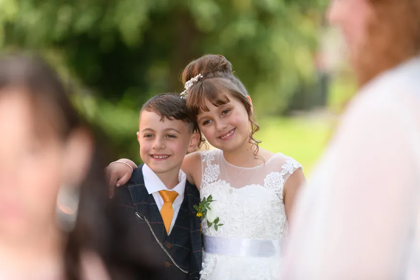 Smiling young boy and girl dressed in formal attire with the girl’s arm around the boy outside in a garden setting.