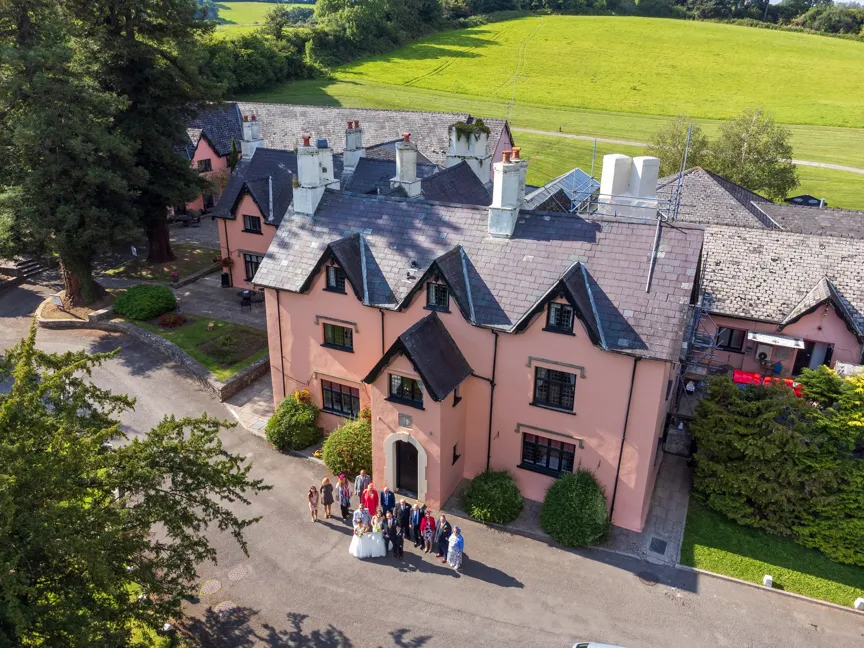 A group of people, including a bride and groom, standing outside a large pink building with slate roofs in a rural setting.