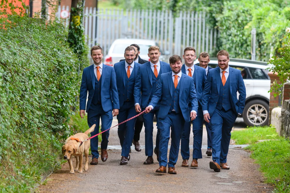 Seven men in blue suits with orange ties walking outdoors on a paved path, one holding a leash walking a yellow Labrador retriever.