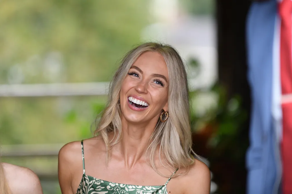 Smiling blonde woman with loose hair wearing a green floral dress and gold hoop earrings.