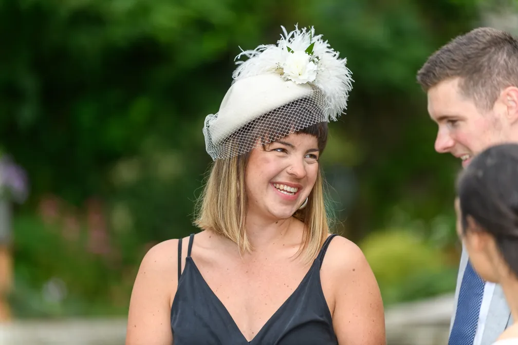 Smiling woman wearing a decorative white fascinator hat with flowers and netting, talking to two people outdoors.