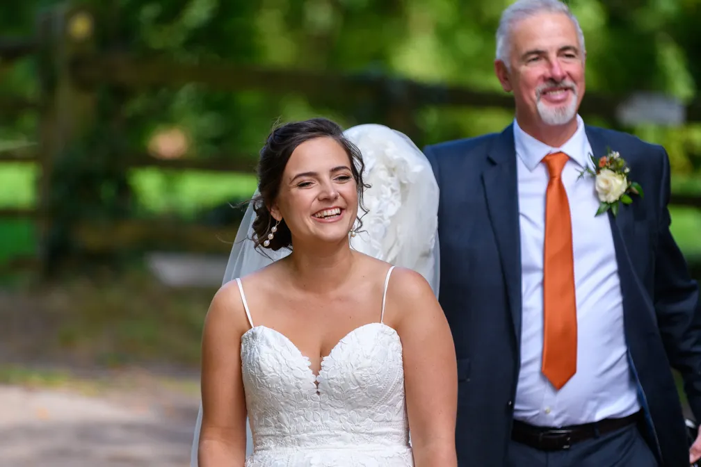 Smiling bride in a white lace wedding dress with a man in a suit and orange tie standing behind her outdoors.