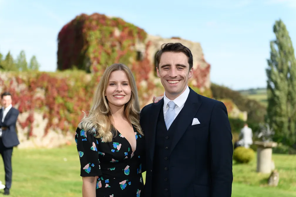 Smiling couple dressed in formal attire standing outdoors with stone ruins and greenery in the background.