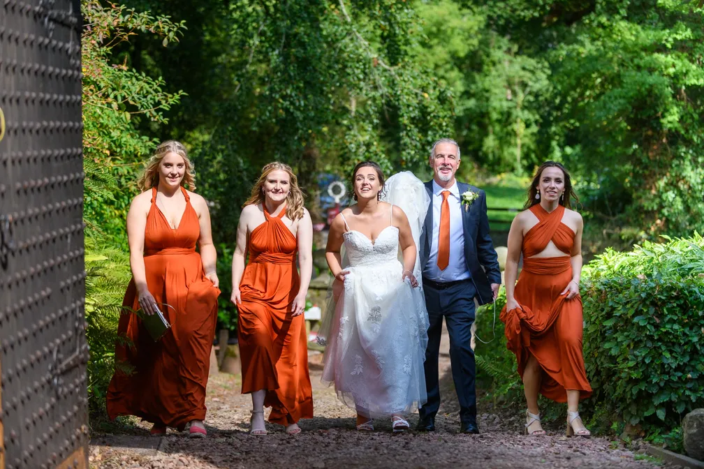 Bride in white wedding dress walking outdoors with three bridesmaids in rust-colored dresses and a man in a dark suit and orange tie.