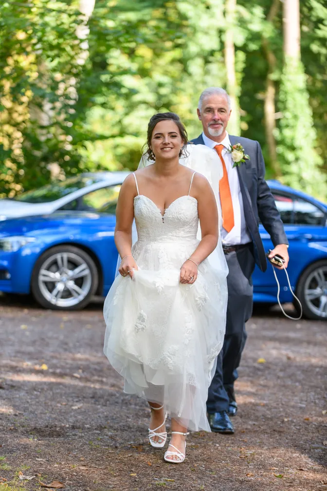 Bride in white wedding dress walking on a path with a man in a suit and orange tie behind her, with a blue car in the background.