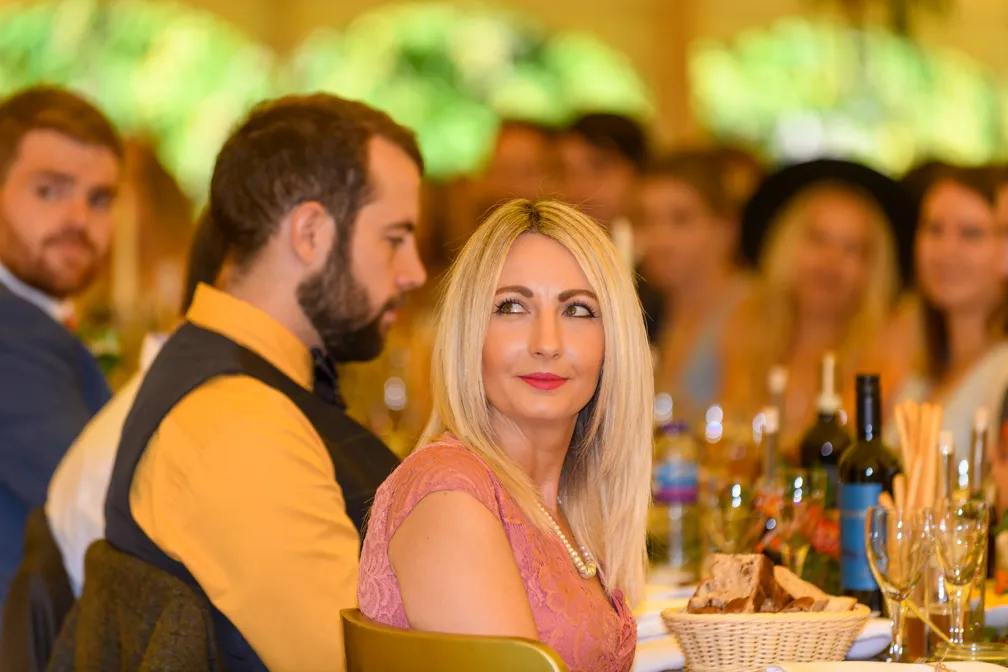 A blonde woman in a pink dress sitting at a table with a man in a yellow shirt and black vest, with other guests and table settings in soft focus in the background.