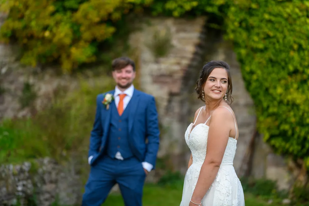 Bride in a white lace wedding dress smiling and looking to the side, with groom in a blue suit and orange tie standing blurred in the background outdoors.
