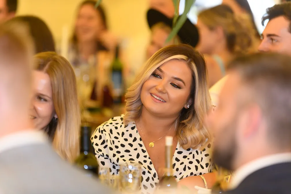 Smiling woman with blonde hair wearing a white and black patterned blouse at a social gathering.