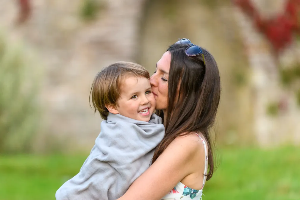 Woman with sunglasses on her head wrapped in a blanket holding and kissing a smiling toddler boy outdoors.