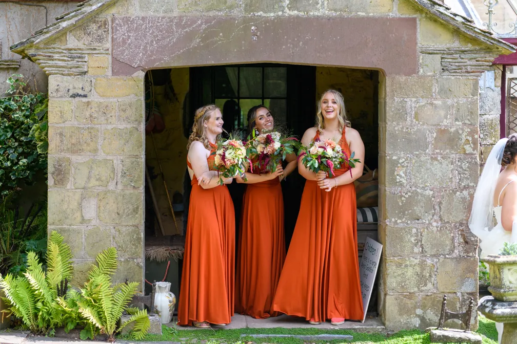 Three bridesmaids in long burnt orange dresses holding bouquets, standing in a stone archway, smiling and talking.
