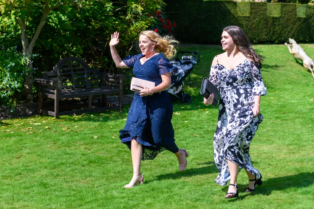 Two women in elegant dresses walking on green grass, one waving and smiling, with a dog jumping in the background.