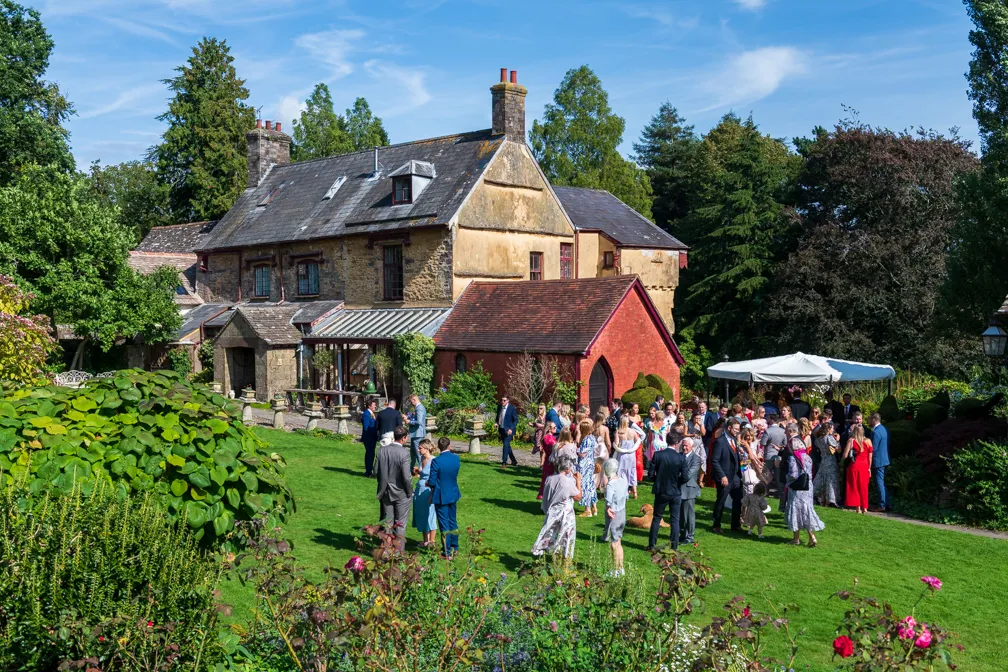 Group of people dressed formally socializing on a sunny lawn in front of a rustic stone and brick building surrounded by trees.