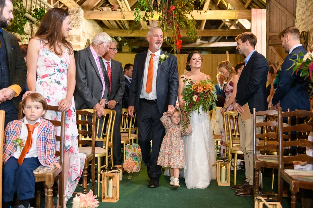 Bride in white dress holding a bouquet walking down aisle with man in suit and child, guests watching in rustic barn setting.