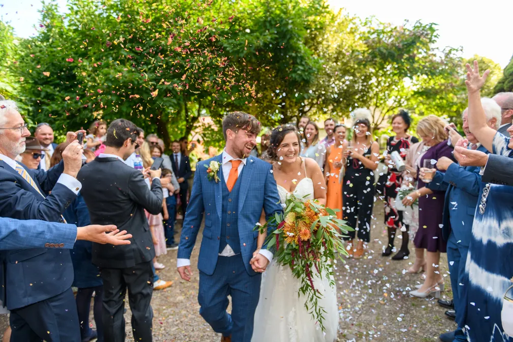 Smiling bride and groom holding hands walking through guests throwing colorful confetti outdoors.