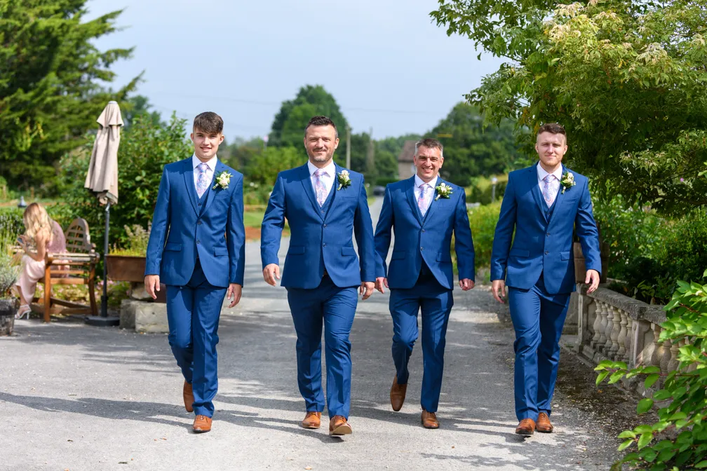 Four men in matching blue suits and brown shoes walking confidently outdoors on a sunny day.