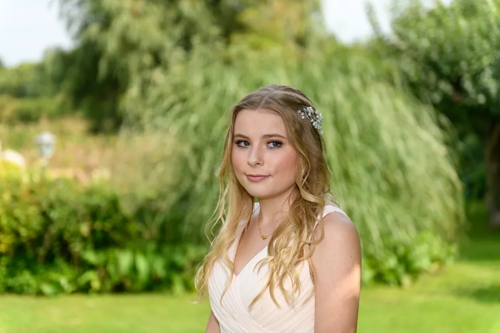 Young woman with long blonde hair wearing a white dress and a hair accessory, standing outdoors with greenery in the background.