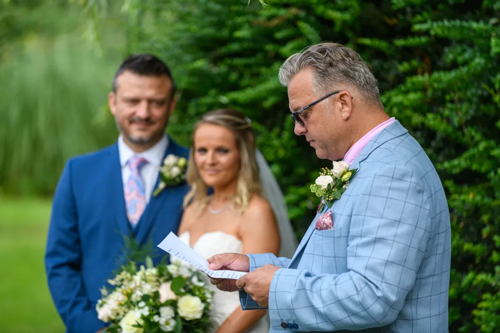 Man in a light blue checked suit reading from a paper with a bride and groom standing behind him outdoors.
