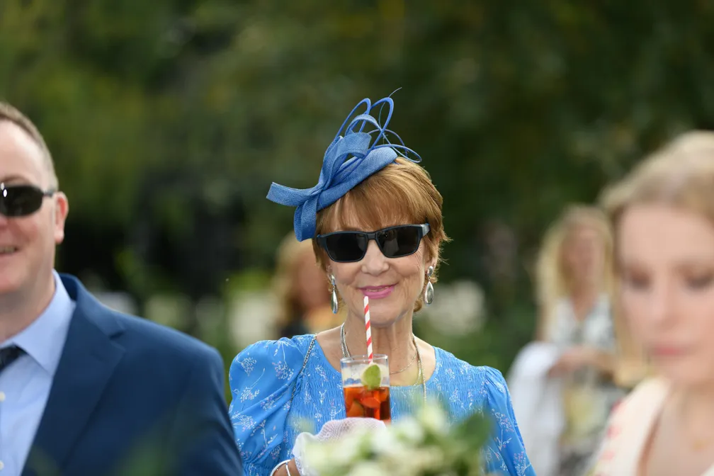 Woman in a blue dress and decorative blue fascinator wearing sunglasses and holding a drink with a red and white striped straw.