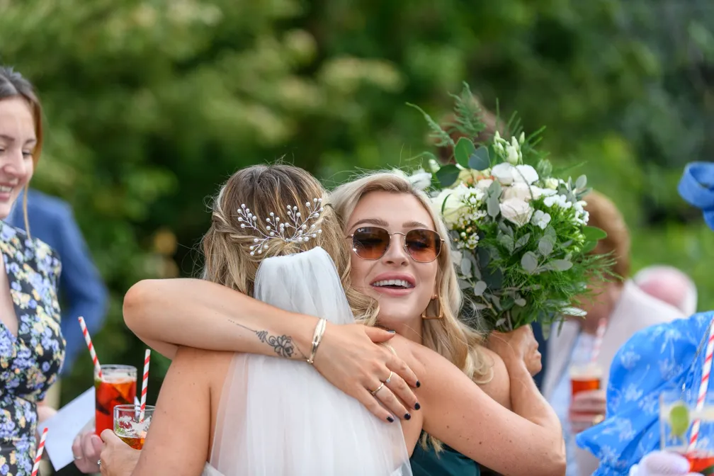Blonde woman wearing sunglasses hugging a bride holding a bouquet of white and green flowers at an outdoor event.