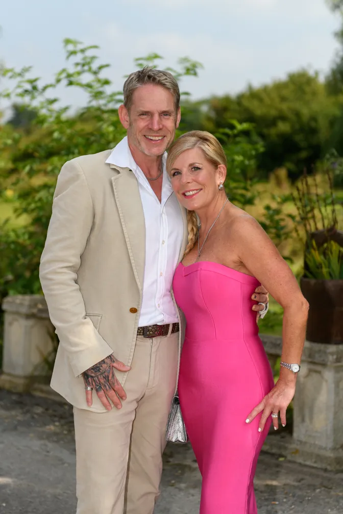 Smiling couple posing outdoors; man in a beige suit with hand tattoo and woman in a strapless pink dress with braided hair.