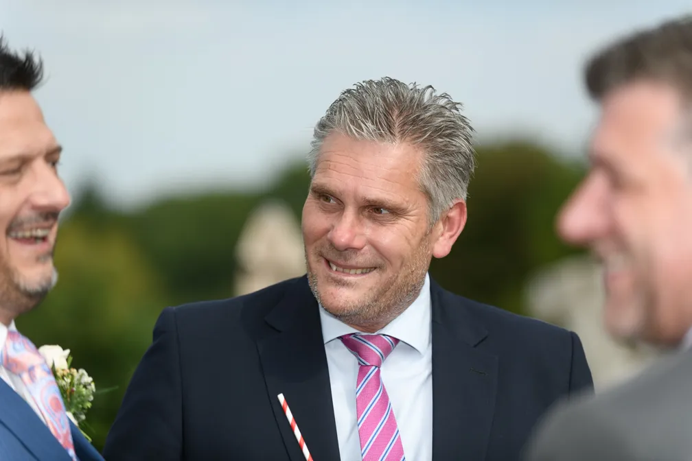 Three men in suits smiling and talking outdoors, with the man in the center wearing a pink striped tie.